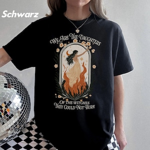 Può includere: T-shirt nera con la scritta "We Are The Daughters Of The Witches They Could Not Burn". La grafica presenta una donna in un abito fluente, circondata da fiamme e elementi floreali. La parola "Schwarz" è in alto a sinistra.