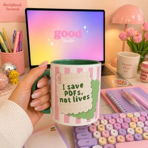 Könnte beinhalten: Ein weißer Keramikbecher mit grünem Innenraum und Henkel, verziert mit rosa-weißen vertikalen Streifen. Eine Grafik eines karierten Notizzettels mit einer roten Büroklammer zeigt den grünen Text: I save PDFS, not lives. Der Becher wird gehalten.