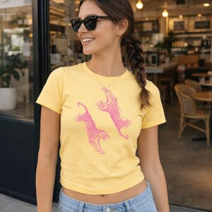 Könnte beinhalten: Ein gelbes T-Shirt mit zwei rosa Tiger-Illustrationen. Das kurzärmelige Oberteil im lässigen Stil ist aus einem weichen Material gefertigt. Die Tiger sind in einer dynamischen Pose dargestellt, was dem Design eine spielerische Note verleiht.