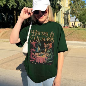May include: Dark green t-shirt with the text "HOUSES & HUMANS" in a decorative font. The graphic depicts dragons playing cards around a table. The dragons are in shades of red, blue, and green.