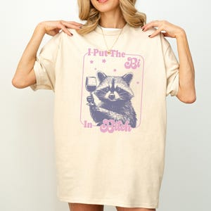 May include: Cream-colored t-shirt featuring a purple graphic of a raccoon holding a wine glass. The text "I Put The Bi In Bitch" is above and below the raccoon illustration. The shirt has short sleeves and a relaxed fit.