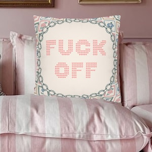 Könnte beinhalten: Ein dekoratives Kissen mit den Worten "FUCK OFF" in Rosa auf cremefarbenem Hintergrund gestickt. Das Kissen hat einen floralen und geometrischen Bordürenrand in Blau und Rosa und liegt auf einem gestreiften rosa-weißen Kissen.