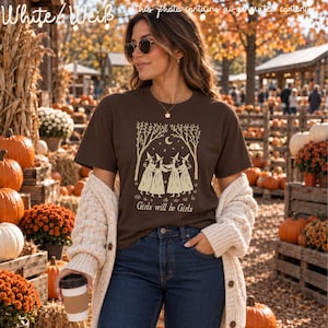 Op de afbeelding: Een donkerbruin T-shirt met korte mouwen en een cr&egrave;mekleurige afbeelding van vier heksen in jurken die elkaars hand vasthouden in een cirkel onder bomen en een maansikkel, met de tekst Girls will be Girls onder de illustratie.