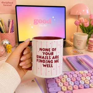 Könnte beinhalten: Ein weißer Keramikbecher mit dunkelrotem Innenraum und Henkel, verziert mit vertikalen rosa Streifen, kleinen Herzen und dem Text 'NONE OF YOUR EMAILS ARE FINDING ME WELL' in dunkelroter Schrift auf der Vorderseite des Bechers.