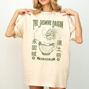 Può includere: T-shirt color crema con grafica verde di un drago che emerge da una tazza da tè, con la scritta "THE JASMINE DRAGON" sopra e caratteri cinesi su entrambi i lati. Il design include la scritta "BA SING SE" su uno stendardo.