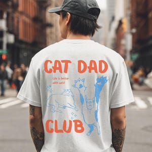 Könnte beinhalten: Hellgraues T-Shirt mit den Worten "CAT DAD CLUB" in Orange und dem Satz "Life is better with cats!" in kleinerer Schrift. Das Shirt zeigt blaue Katzenillustrationen.