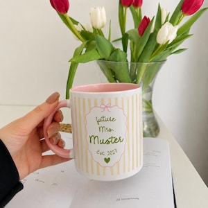 Könnte beinhalten: Eine Keramiktasse mit hellrosa Innenseite und Henkel, verziert mit vertikalen gelben Streifen auf der Außenseite. Die Vorderseite zeigt einen dekorativen gewellten Rahmen mit dem Text "future Mrs. Muster Est. 2027" in Grün, ergänzt durch ein Herz.