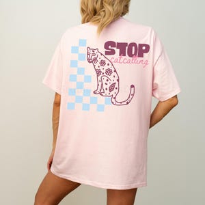 May include: Light pink t-shirt with a graphic of a floral leopard and the text "STOP catcalling". The design includes a blue and white checkered pattern. The shirt is a relaxed fit.