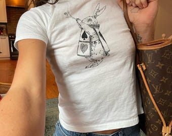 May include: White t-shirt featuring a black and white illustration of a rabbit playing a trumpet, with playing card suit symbols on its coat. The shirt is paired with blue jeans. A brown handbag with a monogram pattern is also visible.