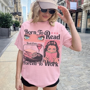 Könnte beinhalten: Rosafarbenes T-Shirt mit dem Text "Born To Read Forced To Work" und Illustrationen von Augen, Büchern und einer lesenden Frau. Das Shirt hat einen Rundhalsausschnitt und kurze Ärmel.