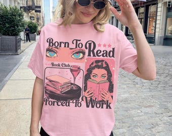 May include: Pink t-shirt with the text "Born To Read Forced To Work" and illustrations of eyes, books, and a woman reading. The shirt has a crew neck and short sleeves.