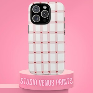 Puede incluir: Una funda de teléfono a cuadros blancos y rosas con detalles de corazones rojos. La funda del teléfono está sobre un pedestal rosa con el texto "STUDIO VENUS PRINTS". La funda tiene un recorte para la cámara y botones laterales negros.