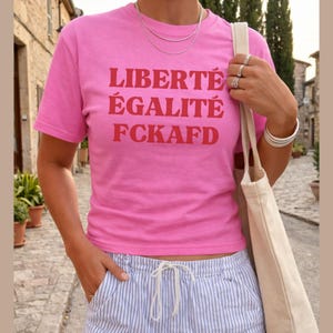 Könnte beinhalten: Ein pinkfarbenes T-Shirt mit dem roten Aufdruck "LIBERTÉ ÉGALITÉ FCKAFD". Dazu werden blau-weiß gestreifte Shorts getragen. Silberner Schmuck, darunter eine Halskette und Armbänder, ist ebenfalls zu sehen.