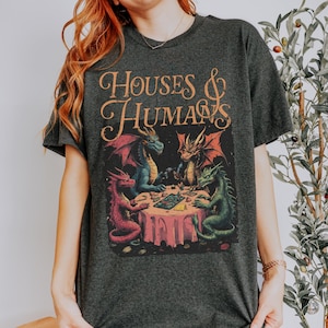 May include: Dark gray t-shirt featuring a fantasy illustration. The graphic depicts four dragons around a table, playing a game. The text "HOUSES & HUMANS" is arched above the scene in orange script.