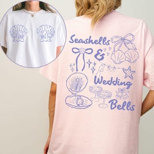 Könnte beinhalten: Ein hellrosa T-Shirt mit dem Schriftzug "Seashells & Wedding Bells" in lila Schrift. Das Shirt zeigt Illustrationen zum Thema Muscheln und Hochzeit in Lila. Ein weißes T-Shirt mit Muschel-Grafiken befindet sich in der oberen linken Ecke.