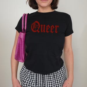 Peut inclure: T-shirt noir avec le mot "Queer" en lettres gothiques rouges. Un sac à main rose vif et texturé est porté sur l'épaule. La personne porte un pantalon à carreaux noir et blanc.