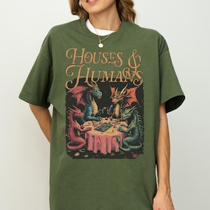 May include: Green t-shirt featuring a fantasy illustration. The graphic shows four dragons around a table with the text "HOUSES & HUMANS" in an ornate font. The dragons are red, blue, and green, with a pink tablecloth.