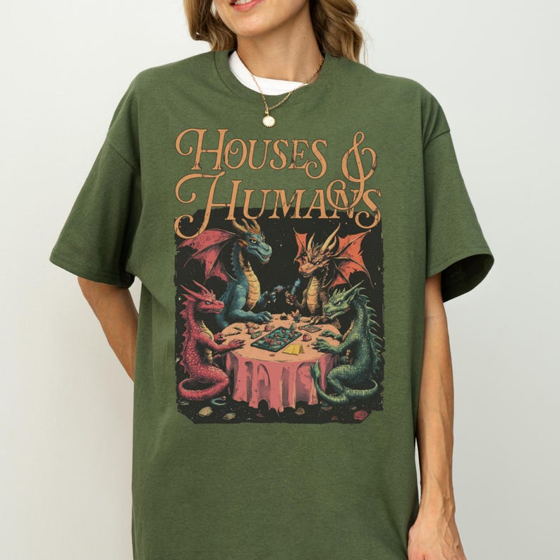 May include: Green t-shirt featuring a fantasy illustration. The graphic shows four dragons around a table with the text "HOUSES & HUMANS" in an ornate font. The dragons are red, blue, and green, with a pink tablecloth.