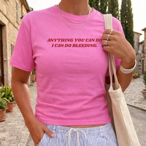 以下が含まれることがあります： 「ANYTHING YOU CAN DO I CAN DO BLEEDING」の赤い文字が入った明るいピンク色のTシャツ。半袖で柔らかい素材のようです。銀色のネックレスと指輪を身につけています。