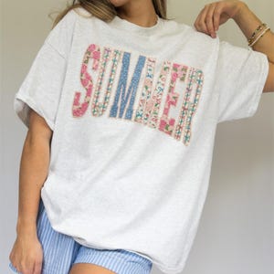 Könnte beinhalten: Ein hellgraues T-Shirt mit dem Wort "SUMMER" in einem Patchwork-Design. Die Buchstaben sind mit Blumen- und Tupfenmustern in Rosa, Blau und Weiß verziert. Das Shirt hat kurze Ärmel.