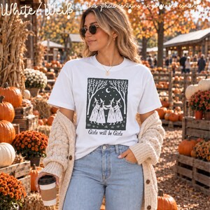 Op de afbeelding: Een wit t-shirt met korte mouwen en een zwarte grafische print van vier figuren met puntmutsen die elkaars hand vasthouden in een cirkel onder een maansikkel en bomen, met de tekst Girls will be Girls onder de illustratie.