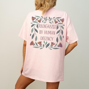 Könnte beinhalten: Hellrosa T-Shirt mit dem Text "RADICALIZED BY HUMAN DECENCY" in einem floralen Kranz-Design. Das Shirt besteht aus einem weichen, bequemen Material.
