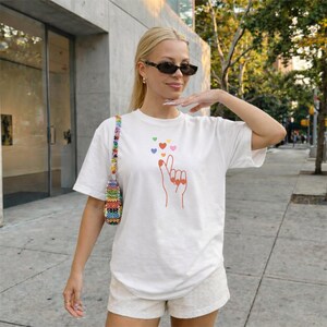 Könnte beinhalten: Weißes T-Shirt mit einer Grafik einer Hand, die ein Herz formt, mit bunten Herzen darüber. Das Outfit umfasst helle Shorts und eine Perlenhandtasche. Die Person trägt eine Sonnenbrille.