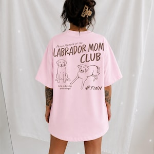 May include: A light pink t-shirt featuring dark brown text that reads Proud Member of the LABRADOR MOM CLUB, Life is better with dogs!, and #FINN. The design includes line art illustrations of two Labrador dogs in a sitting and lying position.