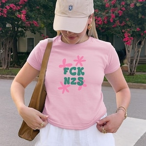 Könnte beinhalten: Rosafarbenes T-Shirt mit dem Text "FCK NZS" in grünen und rosa Blumenmustern. Das kurzärmelige Oberteil ist aus weichem Stoff. Die Person trägt eine beige Baseballkappe und einen weißen Rock.