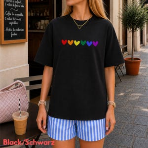 Könnte beinhalten: Schwarzes T-Shirt mit einem Regenbogen-Herz-Design. Das Model trägt blau-weiß gestreifte Shorts. Eine goldene Halskette und Uhr sind sichtbar. Der Text "Black/Schwarz" ist unten.