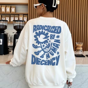 Könnte beinhalten: Ein weißes Sweatshirt mit dem blauen Text "RADICALIZED BY HUMAN DECENCY" und einem Blumendesign mit einer Taube. Das Sweatshirt ist ein lässiger Stil, geeignet für den Alltag.
