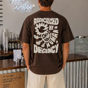 Könnte beinhalten: Braunes T-Shirt mit dem weißen Schriftzug "RADICALIZED BY HUMAN DECENCY" und einem floralen Muster. Die Person trägt beige Shorts. Im Hintergrund ein Café mit Leuchtreklame.