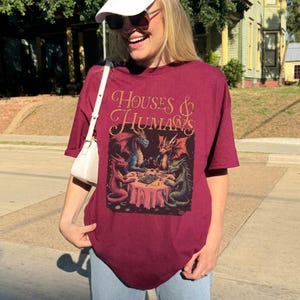 May include: Burgundy t-shirt with the text "HOUSES & HUMANS" in gold script. The graphic depicts four dragons around a table. The dragons are red, blue, and green. The shirt is a casual, short-sleeved style.