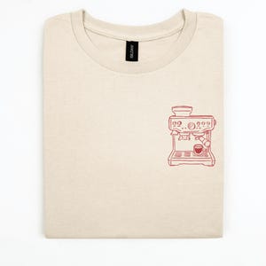 Könnte beinhalten: Ein sandfarbenes T-Shirt mit Rundhalsausschnitt und einer kleinen, roten Strichzeichnung einer Espressomaschine auf der rechten Brustseite. Das T-Shirt hat ein schwarzes Gildan-Markenetikett am inneren Halsausschnitt.
