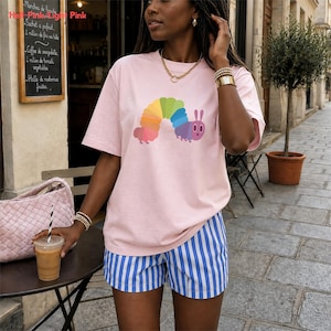 Könnte beinhalten: Hellrosa T-Shirt mit einem bunten Raupen-Design. Dazu blaue und weiße gestreifte Shorts. Die Person trägt Goldschmuck und hält ein Getränk mit einem Strohhalm.