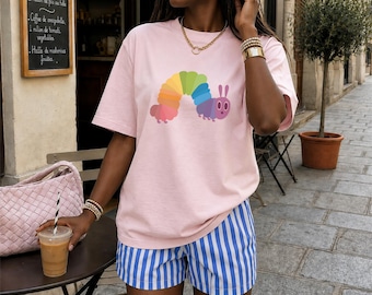 May include: A light pink t-shirt featuring a colorful caterpillar design. The shirt is paired with blue and white striped shorts. The person is holding a drink with a straw and wearing gold jewelry.