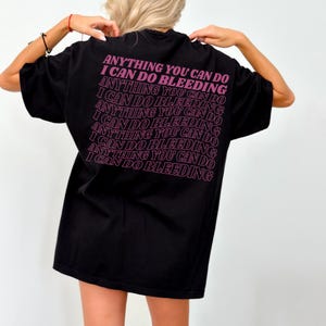 Könnte beinhalten: Schwarzes T-Shirt mit dem pinkfarbenen Text "ANYTHING YOU CAN DO I CAN DO BLEEDING" auf dem Rücken, mehrfach wiederholt. Das Shirt hat einen lässigen Stil.