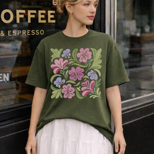 Könnte beinhalten: Olivgrünes T-Shirt mit einem Blumendesign aus rosa, lila und blauen Blumen und grünen Blättern. Das Shirt hat einen Rundhalsausschnitt und kurze Ärmel. Das Design befindet sich mittig auf der Vorderseite.