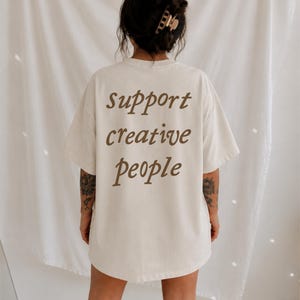以下が含まれることがあります： 背面に筆記体で茶色の文字「support creative people」がプリントされた、オフホワイトの半袖Tシャツ。ゆったりとしたリラックスフィットのシルエットで、クルーネックのデザインが背中の上部を覆っています。