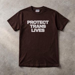 Könnte beinhalten: Ein braunes T-Shirt mit dem weißen Schriftzug "PROTECT TRANS LIVES". Das kurzärmelige T-Shirt hat einen Rundhalsausschnitt und ist aus einem weichen Stoff gefertigt. Das Gildan-Markenetikett ist am Halsausschnitt sichtbar. Ein lässiges Kleidungsstück.