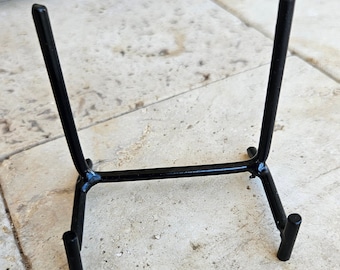May include: A black metal display stand featuring two vertical support arms connected by a horizontal base bar, with two small front-facing hooks designed to hold items securely in an upright position for viewing or presentation purposes.