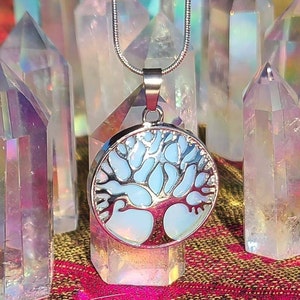 Puede incluir: Un collar de cadena de plata con un colgante redondo que presenta un diseño de árbol de la vida. El colgante está hecho de ópalo con un acabado iridiscente azul y blanco. El collar se muestra sobre una cama de cristales iridiscentes.