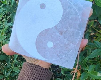 Könnte beinhalten: Ein weißer, quadratischer, polierter Stein mit einem Yin-Yang-Symbol, das in die Oberfläche graviert ist. Der Stein wird von einer Hand vor einem grünen, belaubten Hintergrund gehalten.