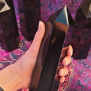 May include: A collection of six black obsidian crystal points, with one point being held in a hand. The points are smooth and polished, with a deep black color.