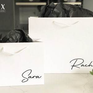 May include: Two white gift bags with black handles and black script names "Sara" and "Rachel" printed on the front.