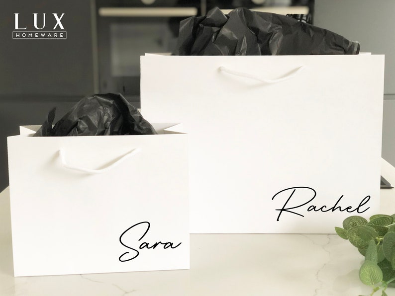 May include: Two white gift bags with black handles and black script names "Sara" and "Rachel" printed on the front.