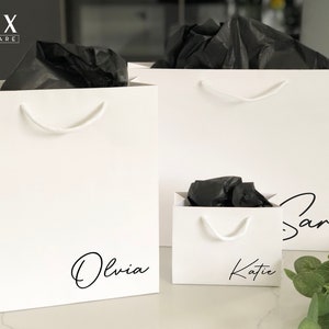 May include: Three white gift bags with black tissue paper inside. The bags are personalised with the names "Olivia", "Katie", and "Sara" in black script.