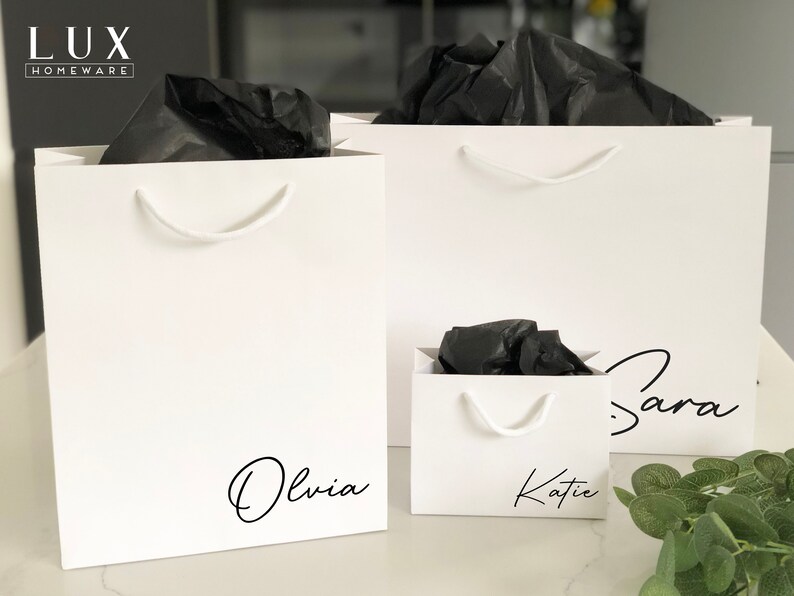 May include: Three white gift bags with black tissue paper inside. The bags are personalised with the names "Olivia", "Katie", and "Sara" in black script.