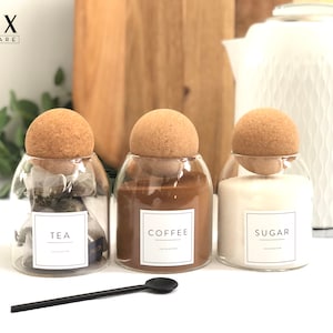 May include: Three glass jars with cork lids and labels that read "Tea", "Coffee", and "Sugar". The jars are filled with tea bags, coffee grounds, and sugar. The jars are on a white surface.