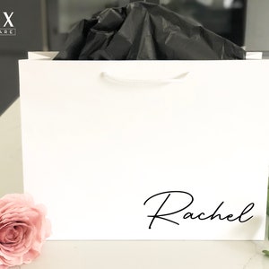 May include: White gift bag with a white rope handle and the name "Rachel" printed in black cursive on the front.